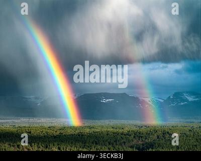 Luftaufnahme eines Doppelregenbogens, der sich über einen dichten grünen Wald und schneebedeckte Berge unter einem dramatischen stürmischen Himmel erstreckt Schottland, Großbritannien. Stockfoto