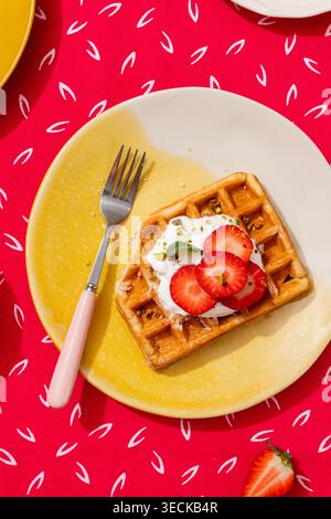 Eine köstliche Waffel mit frisch geschnittenen Erdbeeren und cremigem Ricotta, serviert auf einem gelben Teller mit rosafarbenem Griff, vor einem lebendigen Teller Stockfoto