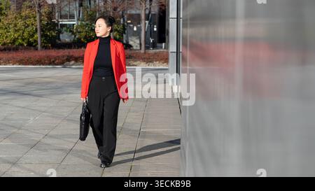 Eine Frau mittleren Alters in einem stilvollen roten Blazer und einer schwarzen Hose spaziert selbstbewusst durch ein modernes Stadtgebiet. Der klare, sonnige Tag bringt ihre Professi Stockfoto