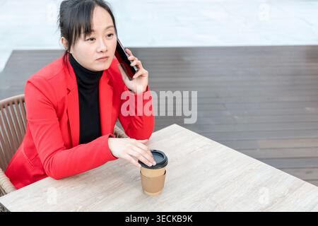 Professionelle Frau mittleren Alters in einem roten Blazer, saß draußen an einem Holztisch mit einem Kaffee zum Mitnehmen, telefonierte, konzentriert und aufmerksam Stockfoto