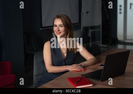 Selbstbewusste junge Geschäftsfrau sitzt an einem Konferenztisch, lächelt, während sie in einem modernen Büro-Besprechungsraum mit Notebook und Smartph an einem Laptop arbeitet Stockfoto
