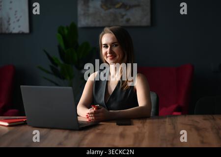 Selbstbewusste junge Geschäftsfrau, die an einem hölzernen Schreibtisch sitzt, lächelt, während sie in einem modernen Büro an einem Laptop arbeitet, und zeigt Professionalität, Konzentration und Sorgfalt Stockfoto