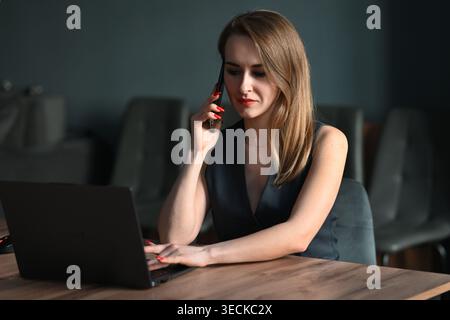 Eine zielgerichtete Geschäftsfrau, die an einem Laptop am Tisch arbeitet, während sie mit einem Smartphone spricht, die Routine im Remote-Büro, Multitasking und professionelle Kommunikation zeigt Stockfoto