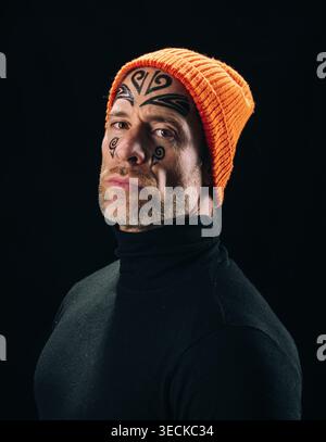 Studio-Porträt eines tätowierten Mannes mit auffälliger Gesichtstinke, orangefarbener Strickmütze und schwarzem Rollkragen, der vor dunklem Hintergrund ernsthaft aussieht Stockfoto