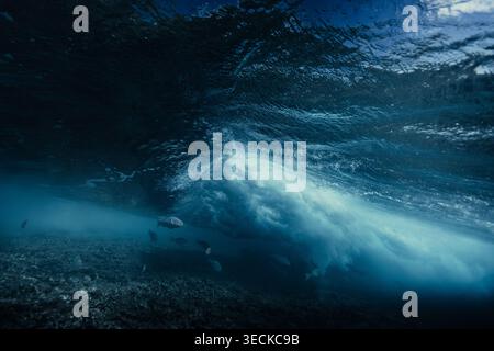 Dramatischer Unterwasserblick auf eine mächtige Meereswelle, die über einem flachen Riff rollt, mit kleinen Fischen und Meerestieren im klaren blauen Wasser in der Nähe eines Maldive Stockfoto