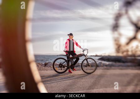 Aktive Frau in roter Jacke steht mit ihrem Elektrofahrrad auf einer ruhigen Küstenstraße und genießt Sonnenuntergangslicht und Meeresbrise während einer entspannten Outdoor-Runde Stockfoto