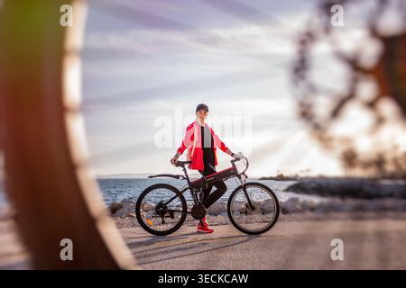 Aktive Frau in roter Jacke steht mit ihrem Elektrofahrrad auf einer ruhigen Küstenstraße und genießt Sonnenuntergangslicht und Meeresbrise während einer entspannten Outdoor-Runde Stockfoto
