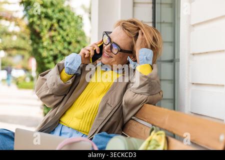 Eine stilvolle Frau mit Brille spricht auf ihrem Smartphone, während sie draußen auf einer Bank sitzt und einen entspannten Lebensstil und eine lebendige Mode in der Sonne präsentiert Stockfoto