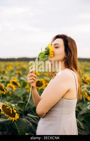 Seitenprofil einer jungen Frau in einem hellen Kleid, die zur goldenen Stunde auf einem blühenden Feld nach Sonnenblume riecht, ruhige Sommerstimmung mit weichem Himmel und natürlichem Kuh Stockfoto