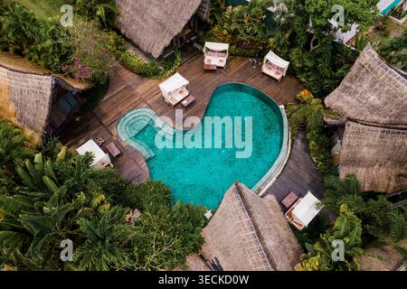 Blick aus der Vogelperspektive auf einen tropischen Resortpool mit Holzterrasse und Cabana-Betten, umgeben von üppigem Dschungel und strohgedeckten Villen in Tegallalang, Bali, Indonesien. Stockfoto