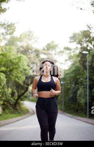 Die junge Frau joggt auf einer ruhigen Parkstraße, die von Bäumen umgeben ist. Sie trägt Sportkleidung und konzentriert sich auf Cardio-Training, Fitness-Routine, Sport und Stockfoto