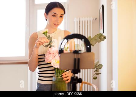 Die junge Frau legt Grün und Blumen in eine Vase, während sie ein Video mit einem Smartphone auf einem Stativ in einem gut beleuchteten Innenraum aufnimmt Stockfoto