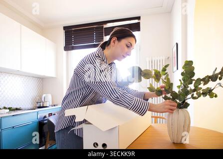 Frau in einem gestreiften Hemd legt Grün in einer weißen Vase auf einen Holztisch in einer modernen Küche mit blauen Schränken und hellem Sonnenlicht Stockfoto