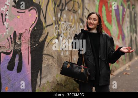 Eine fröhliche junge Frau steht selbstbewusst vor einem farbenfrohen Wandgemälde in einer belebten Stadtallee. Sie trägt eine schicke schwarze Jacke und hält eine Handtasche, strahlend Stockfoto