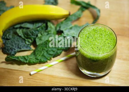 Hochwinkelansicht eines gesunden grünen Smoothies in einem Glas mit gestreiftem Strohhalm. Frische Grünkohl-Blätter und eine gelbe Banane sind im Hintergrund auf einem verschwommen Stockfoto
