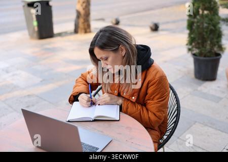 Kreative Momente entfalten sich, wenn eine Frau in ihr Notebook schreibt und die frische Luft in einem lebhaften Café im Freien genießt Stockfoto