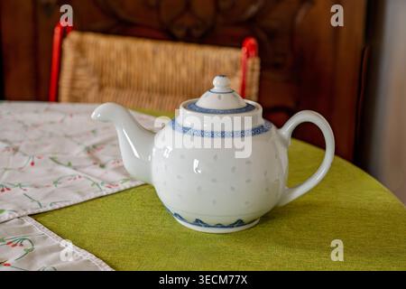 Weiße Keramik-Teekanne mit blauer dekorativer Borte auf einer grünen Tischdecke neben einer Blumenserviette, mit gewebtem Stuhl und geschnitztem Holzhintergrund Stockfoto