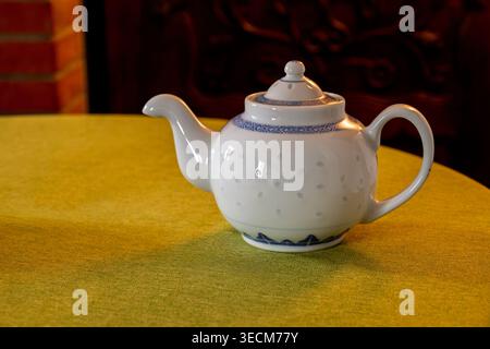 Elegante Teekanne aus weißer Keramik mit blauer dekorativer Borte auf einer grünen Tischdecke, beleuchtet durch warmes Innenlicht mit Holz- und Ziegelbeinfläche Stockfoto