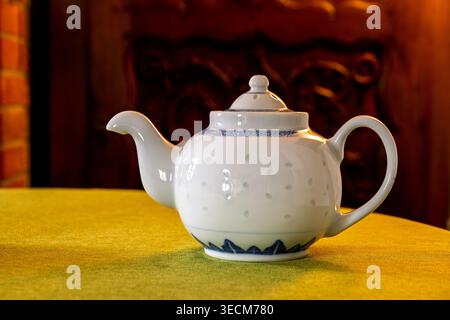 Elegante Teekanne aus weißer Keramik mit blauer dekorativer Borte auf einer grünen Tischdecke, beleuchtet durch warmes Innenlicht mit Holz- und Ziegelbeinfläche Stockfoto