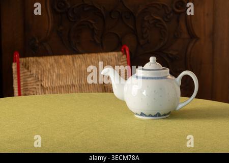 Weiße Keramik-Teekanne mit blauer dekorativer Borte auf einer grünen Tischdecke neben einer Blumenserviette, mit gewebtem Stuhl und geschnitztem Holzhintergrund Stockfoto