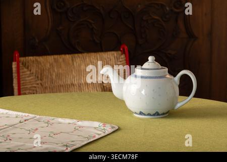 Weiße Keramik-Teekanne mit blauer dekorativer Borte auf einer grünen Tischdecke neben einer Blumenserviette, mit gewebtem Stuhl und geschnitztem Holzhintergrund Stockfoto