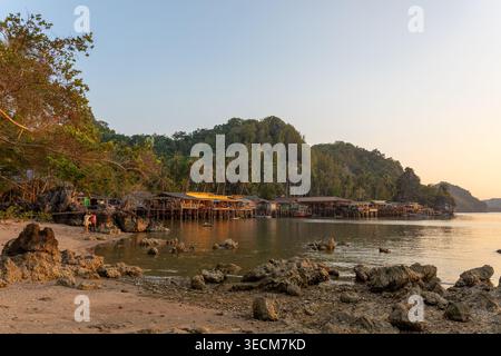 Friedliche, malerische Küstenlandschaft mit felsigem Strand, ruhigem Wasser und traditionellen Holzpfahlhäusern in der Nähe üppiger grüner Berge unter ruhiger Twilig Stockfoto