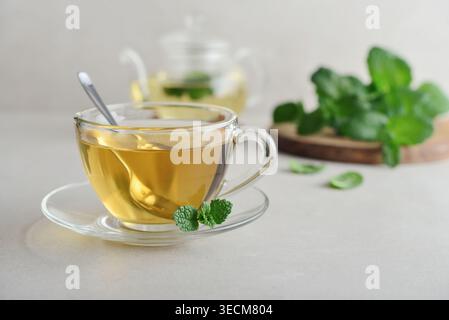 Eine Glasschale mit frischem Minztee auf hellem Hintergrund mit einer Teekanne aus Glas und Minzblättern auf einem Holzbrett im Hintergrund. Konzept des gesunden pflanzlichen d Stockfoto