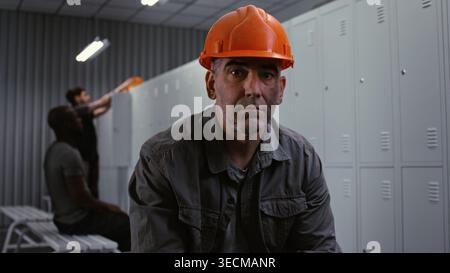 Nahaufnahme eines männlichen Arbeiters mittleren Alters im orangefarbenen Schutzhelm, der die Kamera mit müdem, aber professionellem Ausdruck im Fabrikschließraum ansah. Authentische Geschichte Mit Blauem Kragen. Hochformat. Stockfoto