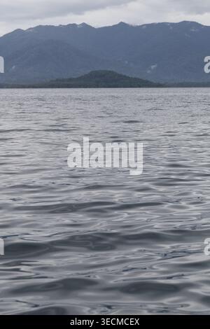 Ruhige Wasseroberfläche mit entferntem Insel- und Berggrund an einem bewölkten Tag. Stockfoto