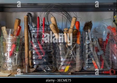 Verschiedene Handwerkzeuge, einschließlich Hämmer, Zangen, Schraubendreher und Metallgeräte, die in wiederverwendeten Kunststoffbehältern auf einem Werkstattregal gelagert werden Stockfoto