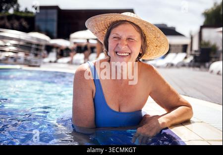 Eine ältere Frau in blauem Badeanzug genießt das Wasser in einem Swimmingpool mit Liegestühlen und einem hellen Himmel, der im Hintergrund sichtbar ist Stockfoto