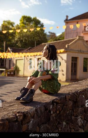 Eine junge Frau mit kurzen Haaren sitzt leise auf einer Steinmauer. Sie schaut sich um das kleine Dorf herum, das mit bunten Fahnen geschmückt ist. Die Sonne scheint hell, Stockfoto