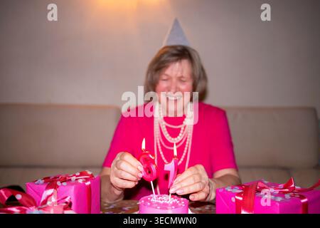 Eine ältere Frau, die ein rosafarbenes Hemd und einen Partyhut trägt, zündet Kerzen auf einer Geburtstagskuche an, mit bunten Geschenken, die auf dem Tisch in einer festlichen Innenumgebung angeordnet sind Stockfoto