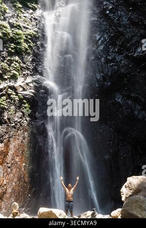 Mann mit erhobenen Armen unter mächtigem Dschungel-Wasserfall, spürt Energie in tropischer Natur Stockfoto