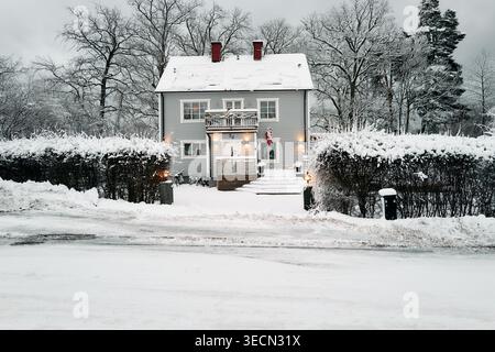 Schnee bedeckt ein gemütliches Haus mit warmen Lichtern. Kiefern stehen hoch hinter schneebedeckten Hecken. Eingangstreppen führen zu einem einladenden Eingang. Nynashamn Stockfoto