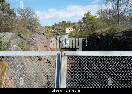 Pendlerzüge kurven durch felsige Schluchten im städtischen Raum. Metallgeländerrahmen im Vordergrund. Nynashamn, Stockholmer County, Schweden, 11. Mai 2025 Stockfoto