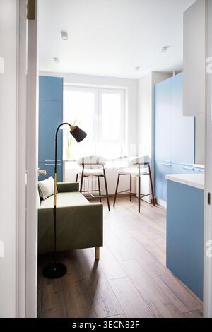 Kleine Küche mit grünem Sofa, blauen Schränken, Holzfußboden und einem Essbereich mit zwei Hockern neben einem großen Fenster Stockfoto