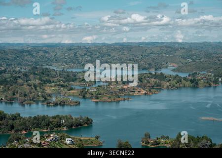 Guatapé Stausee mit zahlreichen grünen Inseln und Halbinseln, umgeben von sanften Hügeln und Wohngebäuden unter einem hellblauen Himmel mit Stockfoto