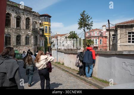 Istanbul, Türkei – 1. April 2026: Menschen gehen eine Kopfsteinpflasterstraße in Balat entlang, vorbei an einem alten Steingebäude und farbenfrohen Wohnhäusern. Architektur Stockfoto