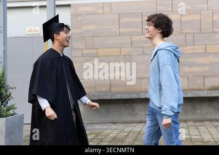 Verschiedene männliche Freunde, einer im Hoodie, feiert den Abschluss im Kleid, Hut mit gelber Quaste im Innenhof Stockfoto