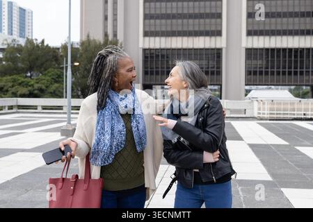 Verschiedene Freundinnen, eine Seniorin, die Arm in Arm am plaza mit roter Tasche und Telefon spricht Stockfoto