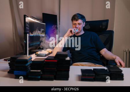 Mann in schwarzem T-Shirt, der elektronische Zigarette während der Arbeit am Computer, umgeben von externen Festplatten, Konzept der Datenspeicherung, Technologie Stockfoto