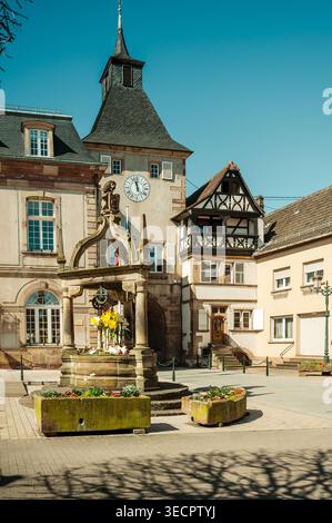 Historischer Stadtplatz mit kunstvollem Brunnen, sonnendurchfluteten Fachwerkfassaden, Steinuhrturm, Blumenkästen, Pflastersteinpflaster, warme Mittagszeit Stockfoto