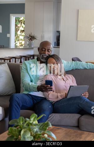 Ein vielseitiges Paar sitzt zu Hause auf einem grauen Sofa, ein Mann zeigt das Smartphone, eine Frau, die ein Tablet hält Stockfoto