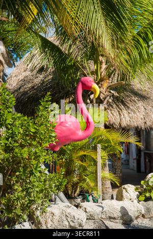 USA, Islamorada, FL - 18. April 2026: Eine große, hellrosa Flamingoskulptur steht in der Nähe von grünen Büschen und einer strohgedeckten Tiki-Hütte an Robbie's Marina. Stockfoto