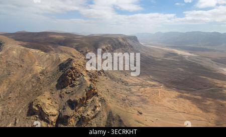 Blick aus der Vogelperspektive auf den zerklüfteten Wüstenberggrat und den Canyon mit felsigen Klippen, kargen Hängen, gewundenen Feldwegen und unbefestigten, weit entfernten Bergketten unter teilweise Clo Stockfoto
