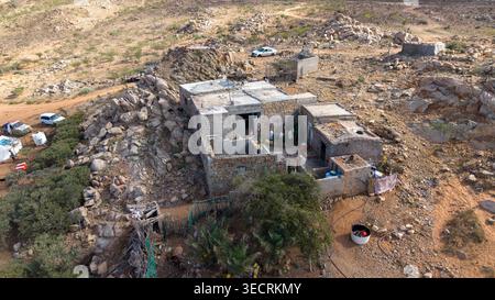 Aus der Vogelperspektive auf ein kleines unvollendetes Steinhaus in felsiger, trockener Hügellandschaft mit verstreuten Felsblöcken, unbefestigten Straßen, wenigen Autos und karger Vegetation. Stockfoto