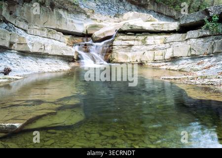 Ein kleiner Fluss bildet einen Wasserfall, der in einen Teich eintaucht, der von Felsen in einem Wald umgeben ist Stockfoto