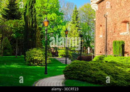 Ein gepflasterter Gang führt durch einen gepflegten Garten mit lebhaften grünen Rasenflächen und Ziersträuchern in Richtung einer alten Backsteinfestungsanlage Stockfoto