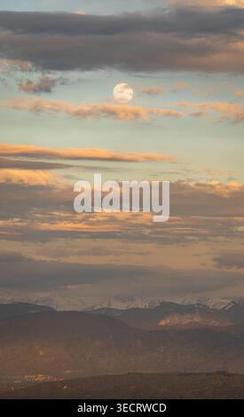 Majestätischer Vollmond scheint in der Abenddämmerung durch verstreute Wolken über ruhigen Bergen. Das weiche, gestreute Licht schafft eine ruhige und mystische Atmosphäre Stockfoto